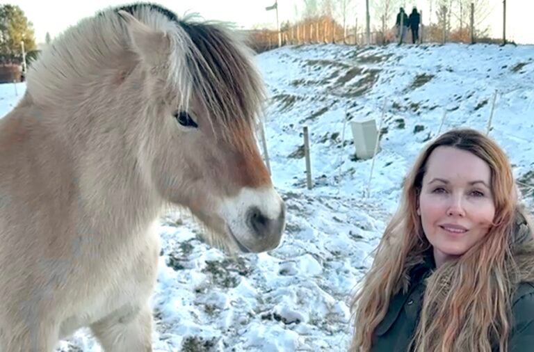 Woman with long blonde hair standing beside a light brown horse in a snowy field
