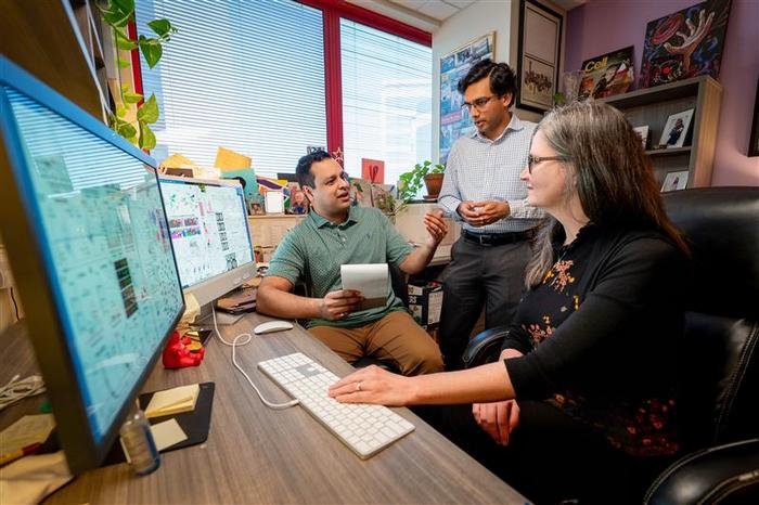 Three coworkers in an office collaborating at a desk a man seated with a tablet another man standing and a woman at a computer