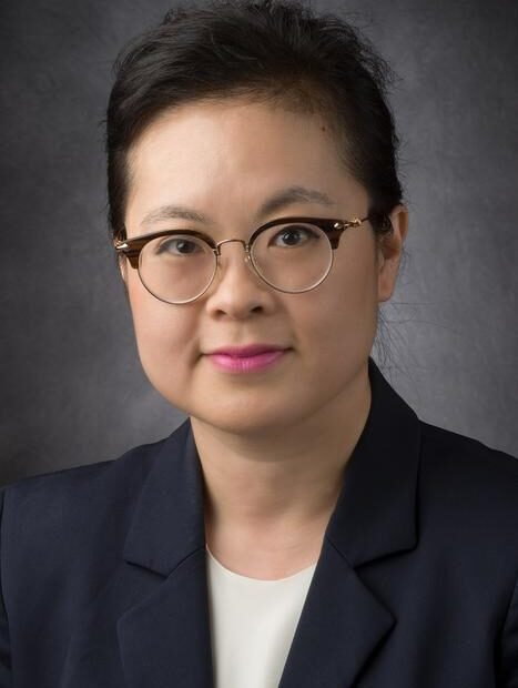 Professional headshot of a woman with dark hair and round glasses wearing a navy blazer and white blouse against a gray backdrop