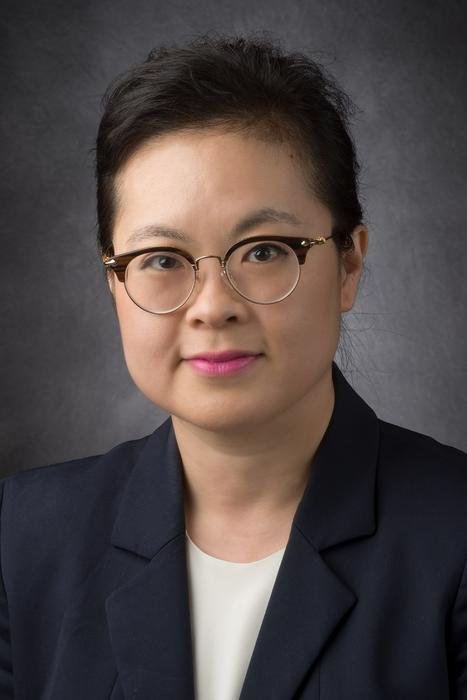 Professional headshot of a woman with dark hair and round glasses wearing a navy blazer and white blouse against a gray backdrop