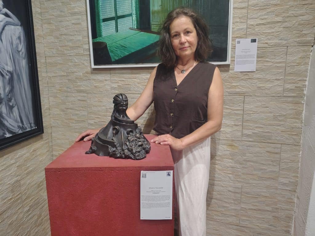 Woman in a brown sleeveless top stands beside a dark sculptural piece on a red pedestal in a gallery setting