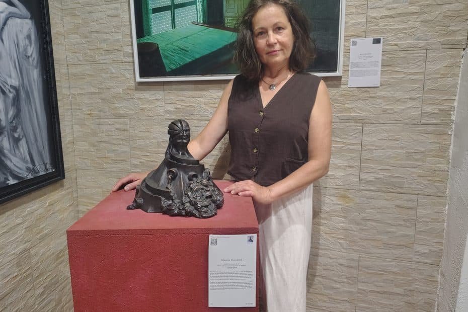 Woman in a brown sleeveless top stands beside a dark sculptural piece on a red pedestal in a gallery setting