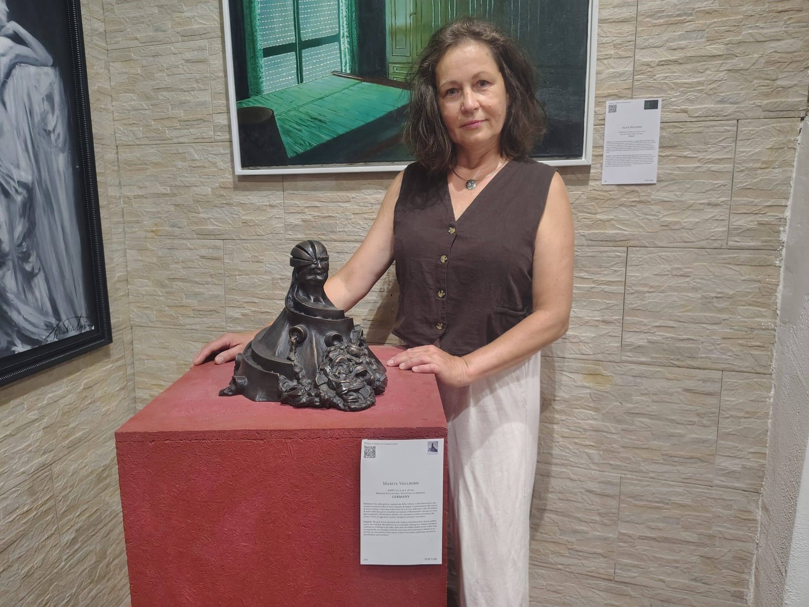 Woman in a brown sleeveless top stands beside a dark sculptural piece on a red pedestal in a gallery setting