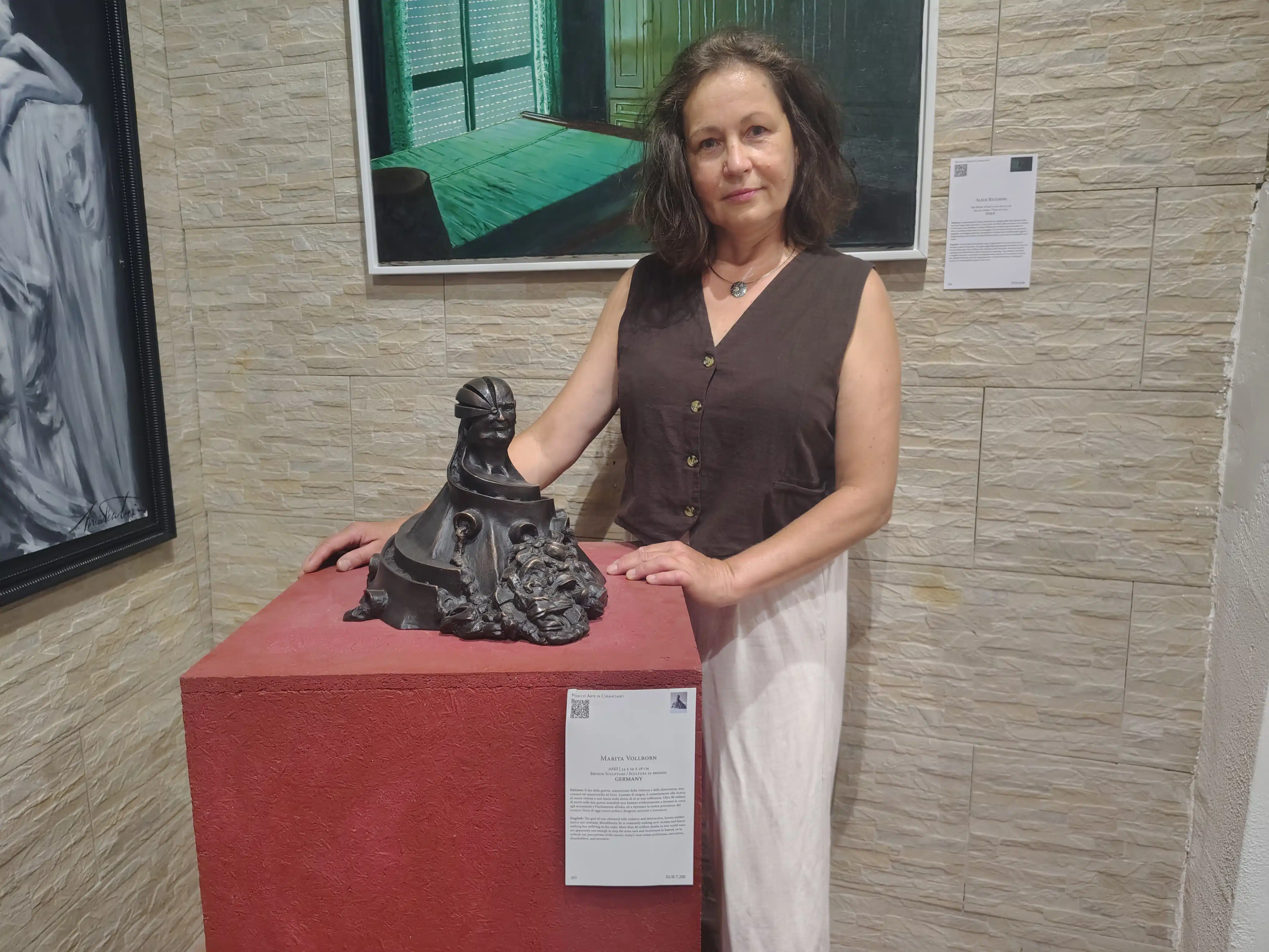 Woman in a brown sleeveless top stands beside a dark sculptural piece on a red pedestal in a gallery setting
