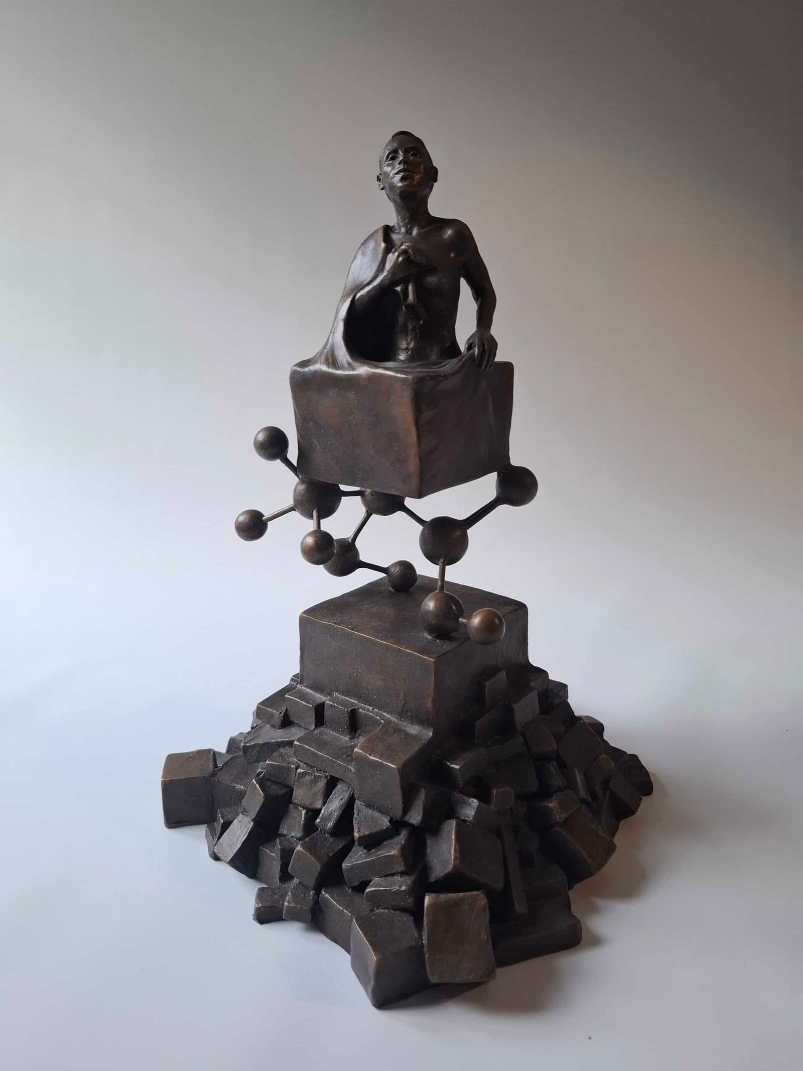 Bronze sculpture of a shirtless man seated on a cube with a molecular like chain of spheres and rods below on a stepped base